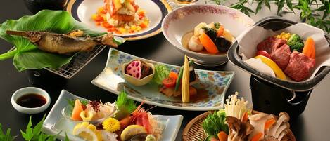 A variety of Japanese dishes including sushi, sashimi, and grilled fish, arranged on a table with a black background and green plants.