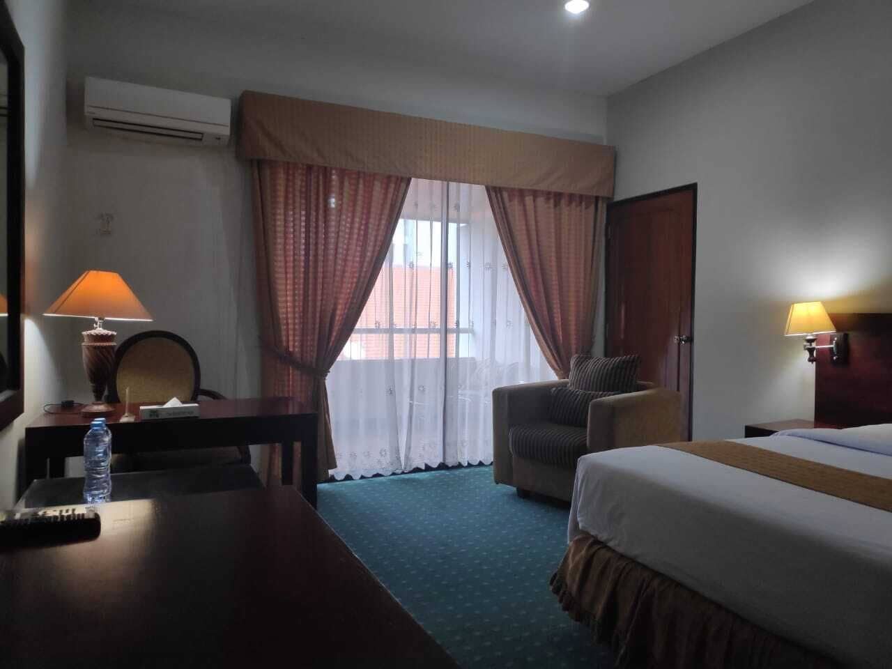 Una habitación de hotel con cama, escritorio, silla, lámpara y ventana con cortinas.