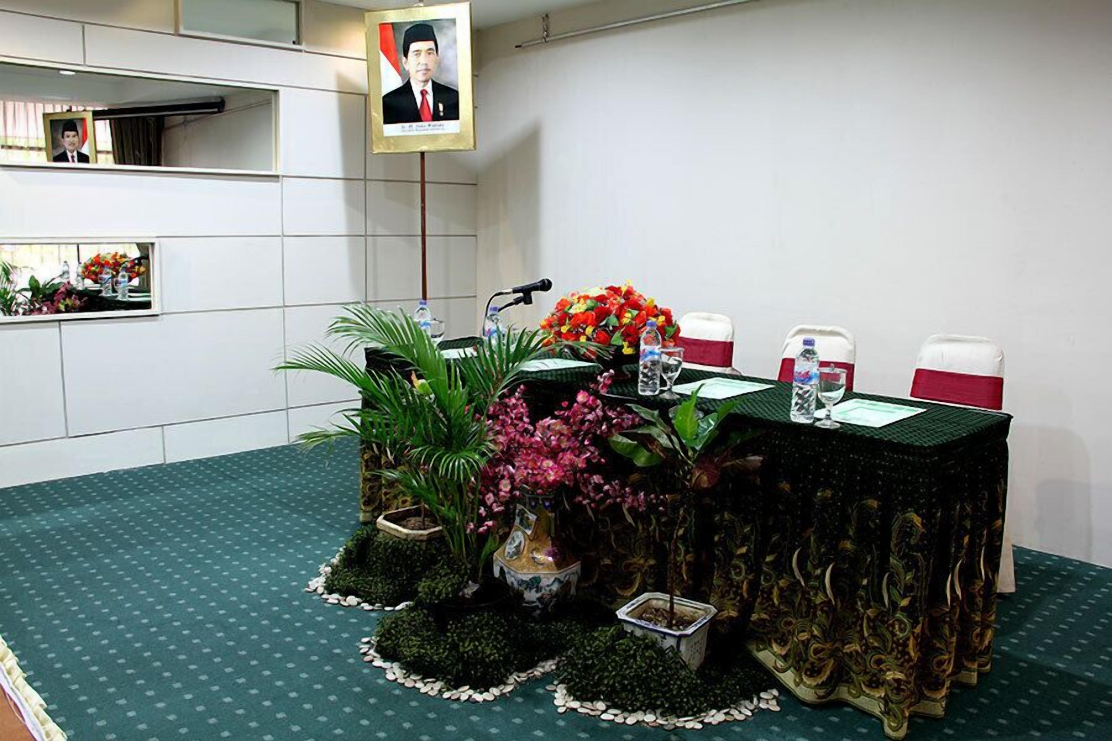 A conference room with a table set for a meeting, surrounded by potted plants and a portrait on the wall.