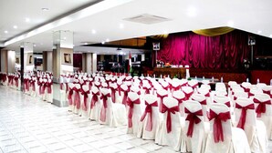 Uma sala de banquetes com fileiras de cadeiras adornadas com fitas vermelhas e brancas, um palco com cortina vermelha e um chão de azulejos.