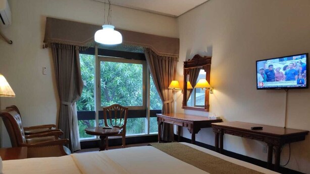 Quarto de hotel com uma cama grande, uma escrivaninha, duas cadeiras, uma televisão e uma janela com cortinas.