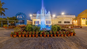 A fountain with water jets, surrounded by potted plants, in an outdoor plaza with a building in the background. - Aapno Ghar Resort (Gurugram)
