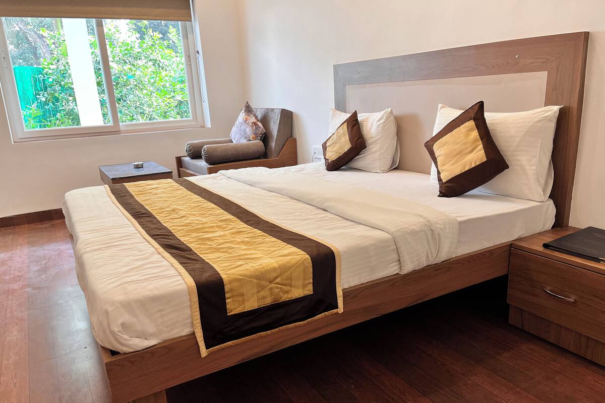 Una cama bien hecha con cabecero de madera, una mesita de noche con un libro y una silla en una habitación con ventana y vistas al exterior con zonas verdes.