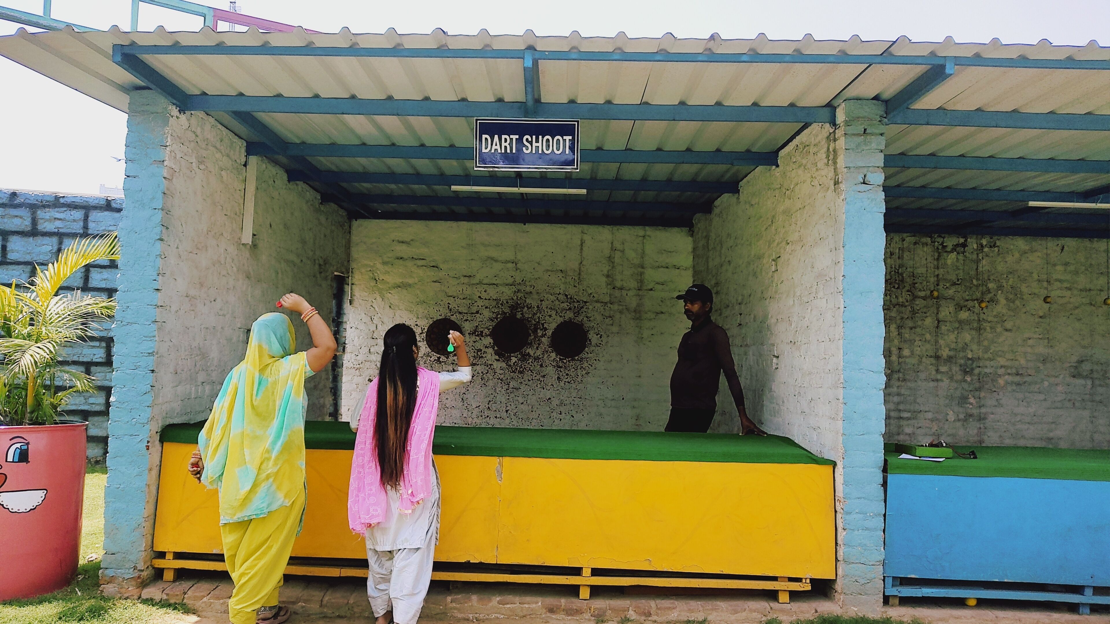 Un tablero de dardos con la leyenda 'DART SHOOT' bajo una zona cubierta con pared de hormigón y suelo azul y amarillo.