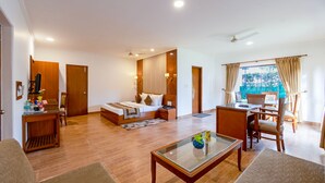 A hotel room with a bed, a sitting area with a table and chairs, a desk, and a view of the outdoors. - Aapno Ghar Resort (Gurugram)