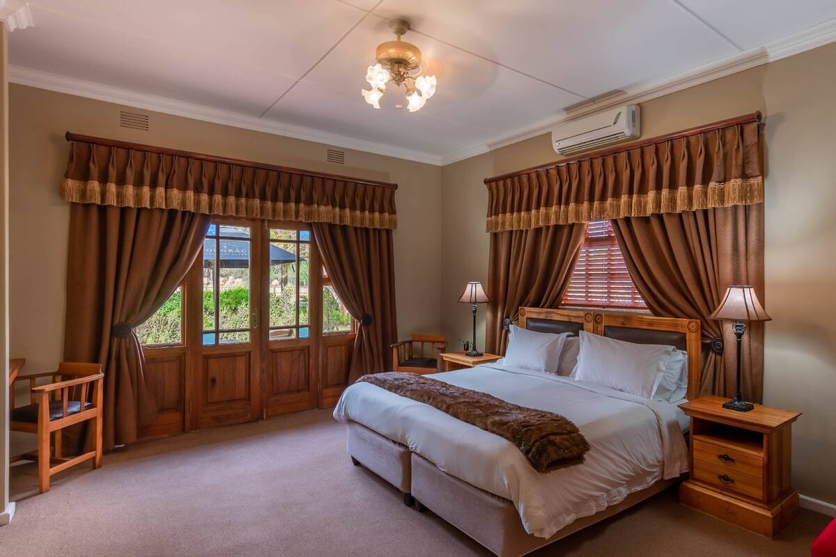 Ein Schlafzimmer mit einem großen Bett, einem hölzernen Kopfteil, zwei Nachttischen mit Lampen und Blick ins Freie durch ein Fenster mit Vorhängen.