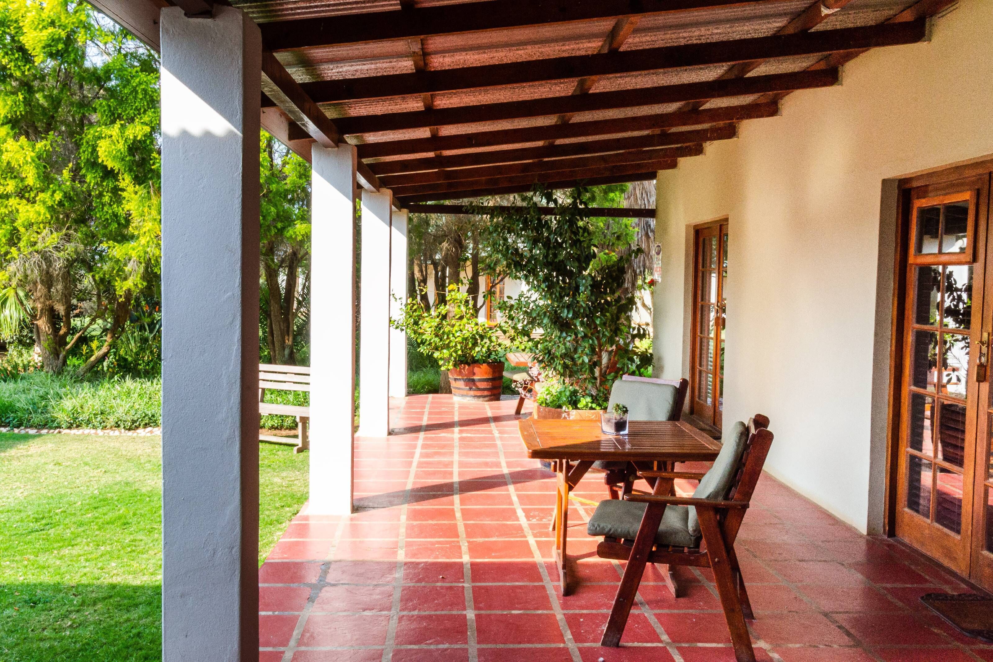 Un porche techado con una mesa y sillas de madera, rodeado de un jardín y árboles.