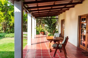 Un porche techado con una mesa y sillas de madera, rodeado de un jardín y árboles.