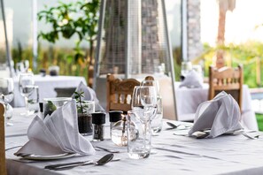 Een eettafel gedekt met witte servetten, glazen en bestek, met zicht op groen en een gebouw op de achtergrond.