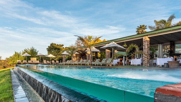 A swimming pool with a waterfall feature, outdoor seating, and a covered area with umbrellas. - Mooiplaas Guesthouse (Oudtshoorn)