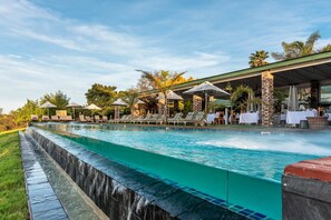 Une piscine agrémentée d’une cascade, un espace extérieur aménagé avec des sièges et une zone couverte avec des parasols.