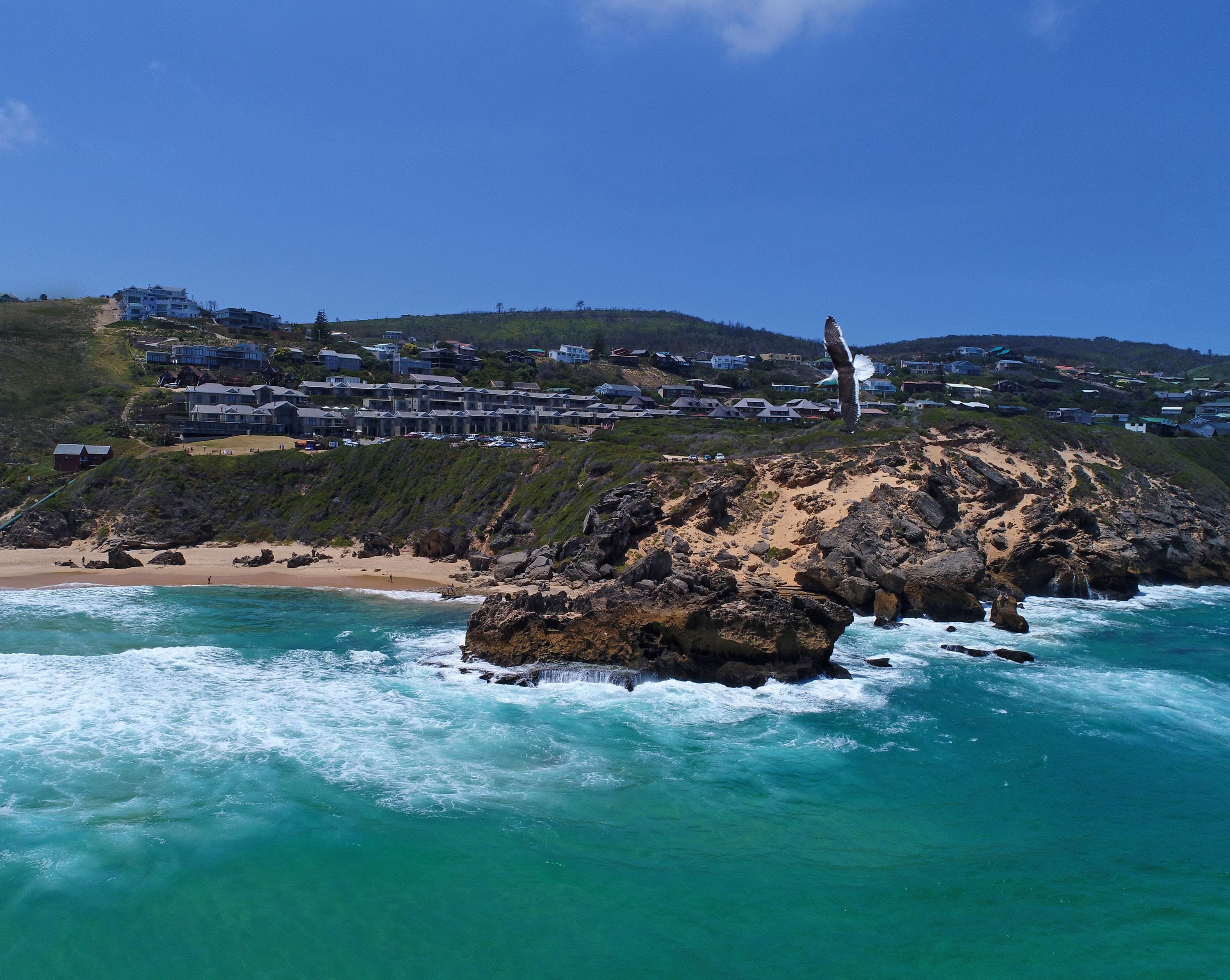 A coastal town with a rocky shoreline, clear turquoise waters, and a view of residential buildings on a hillside.