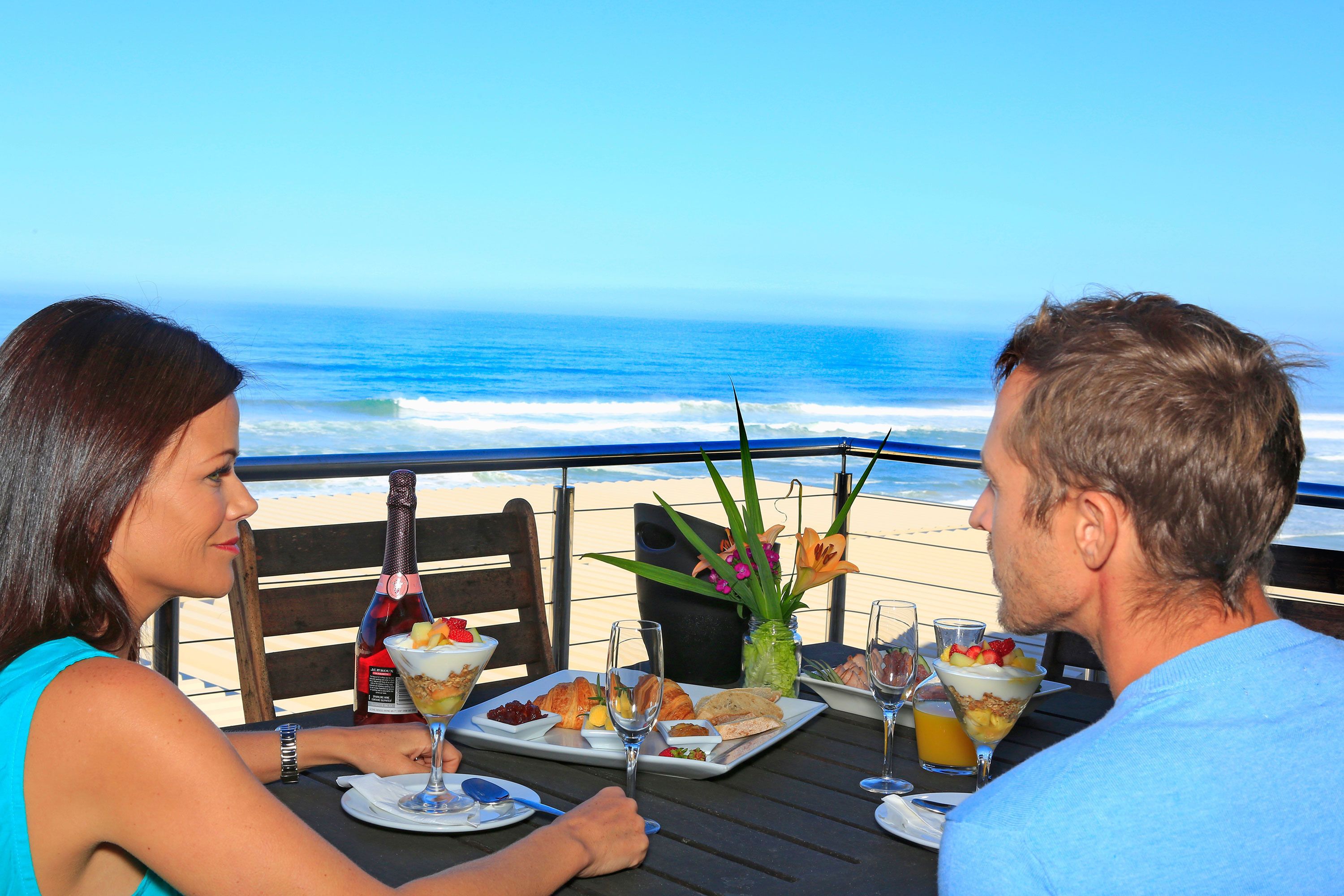 A dining table with food and drinks, overlooking a beach with waves.