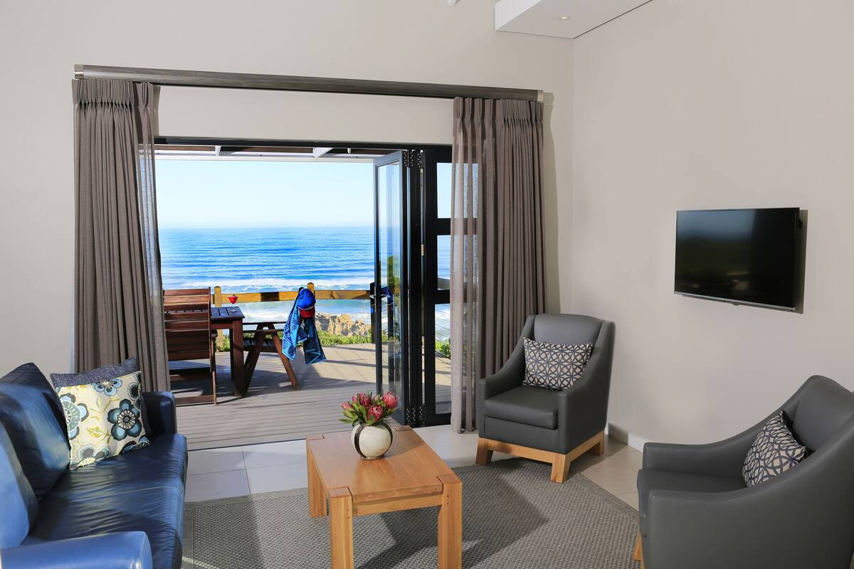 Een kamer met een bank, fauteuil en een tafel met een bloevas. Er is een televisie aan de muur en uitzicht op de oceaan door een glazen deur.