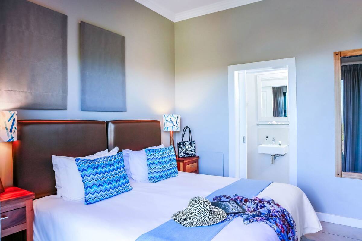Een hotelkamer met een groot bed, een nachtkastje, een lamp, een spiegel, een badkamer met een wastafel en een raam met gordijnen.