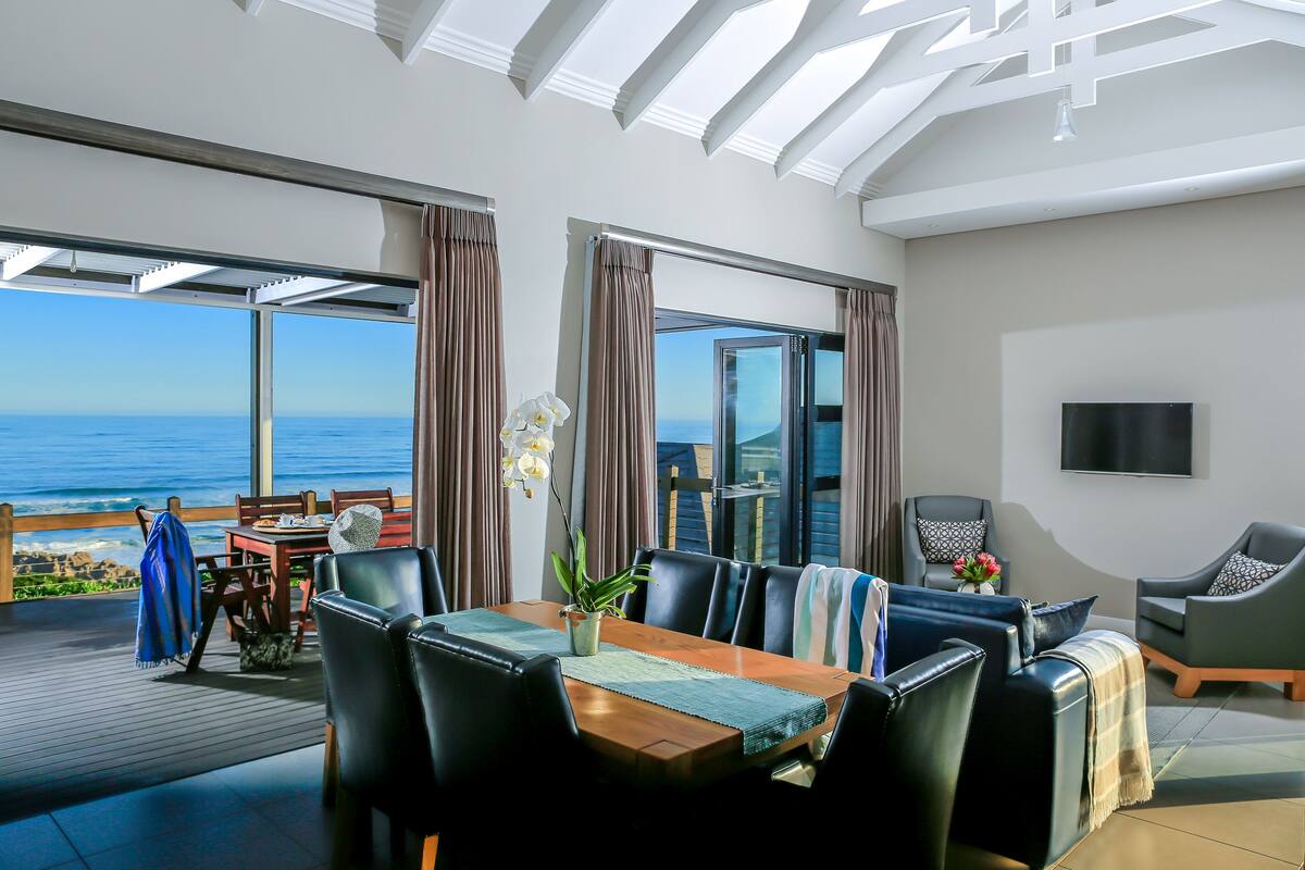Een moderne woonkamer met een grote houten tafel, zwarte leren stoelen en uitzicht op de oceaan.