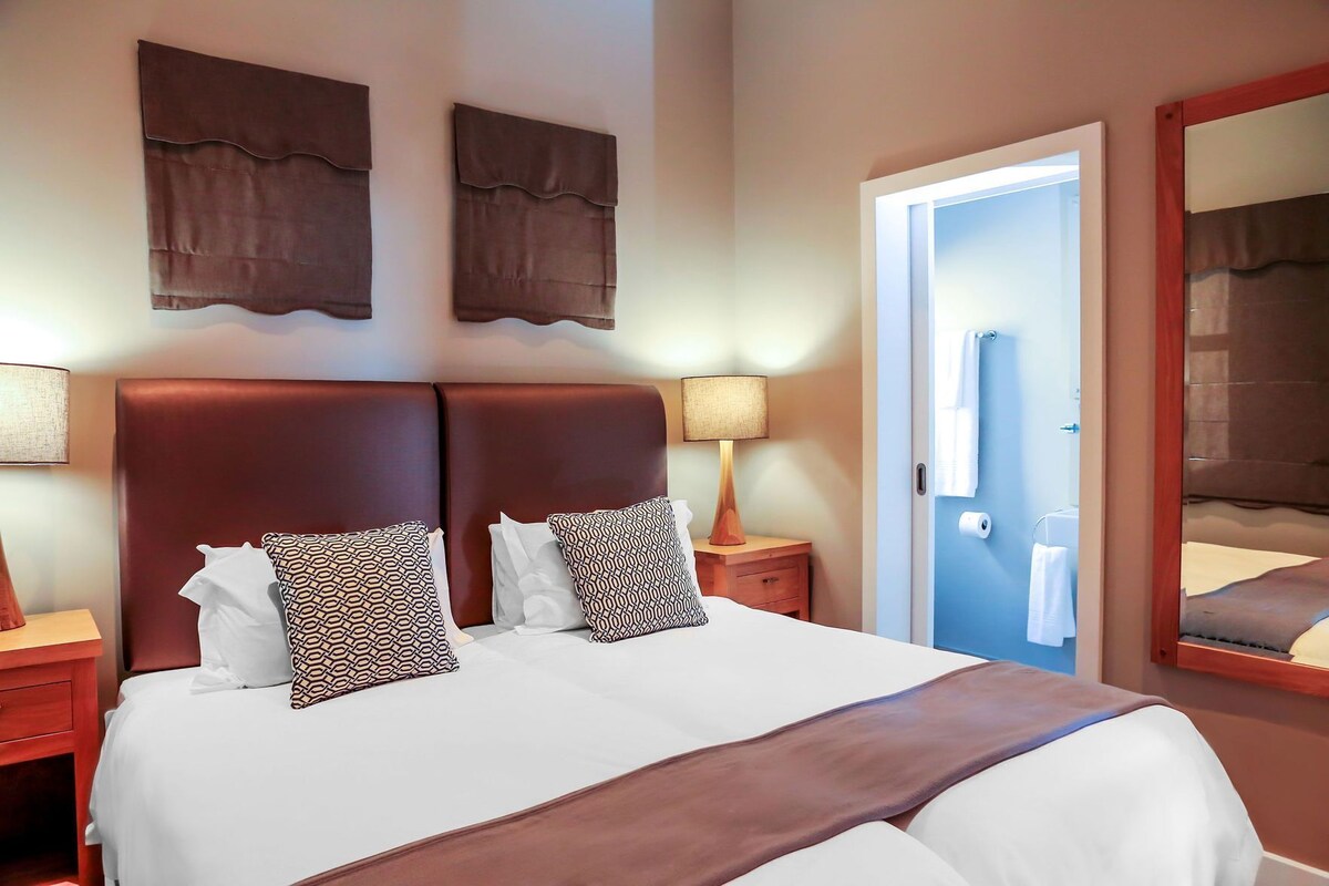 Habitación de hotel con una cama grande, dos mesitas de noche, un tocador, un espejo y una puerta que da a un baño.