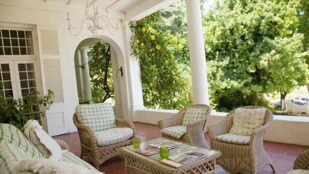 A covered outdoor seating area with wicker furniture and a glass-top table, surrounded by greenery and white walls. - Diemersfontein Wine and Country Estate (Wellington)