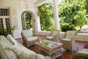 A covered outdoor seating area with wicker furniture and a glass-top table, surrounded by greenery and white walls.