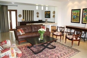Un salon avec un canapé en cuir marron, une table basse et des tapis à motifs. On y trouve également un coin repas avec des chaises en bois et une table. Au fond de la pièce, il y a une cuisine avec des rideaux à rayures.