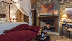 A bedroom with a red velvet sofa, a canopy bed, a stone fireplace, and a painting of a sunset. - Mount Camdeboo Private Game Reserve by NEWMARK (Blue Crane Route)
