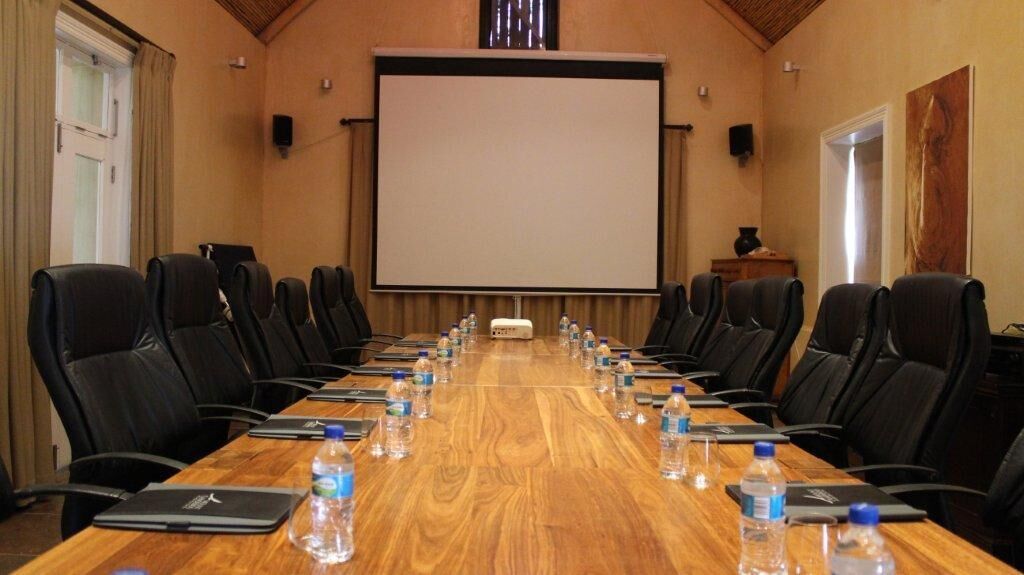 Uma sala de conferências com uma mesa comprida, cadeiras de couro preto, garrafas de água e uma tela de projetor.