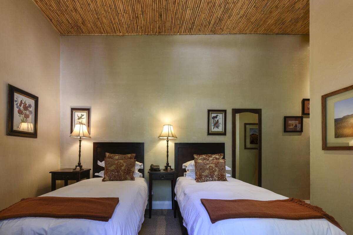 A hotel room with two beds, each with a pillow and a bedside table with a lamp. There are framed pictures on the walls and a mirror on the door.