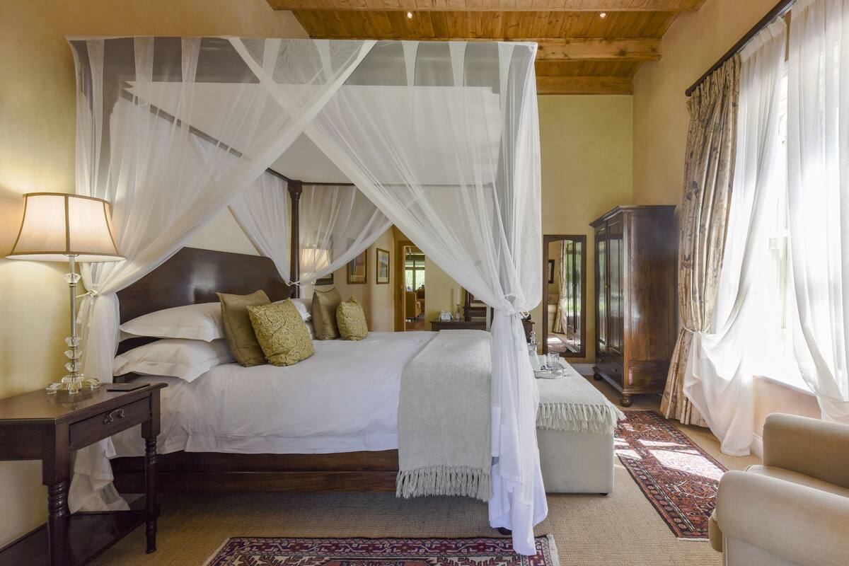 A bedroom with a four-poster bed, a bedside table with a lamp, a wooden wardrobe, and a large window with curtains.