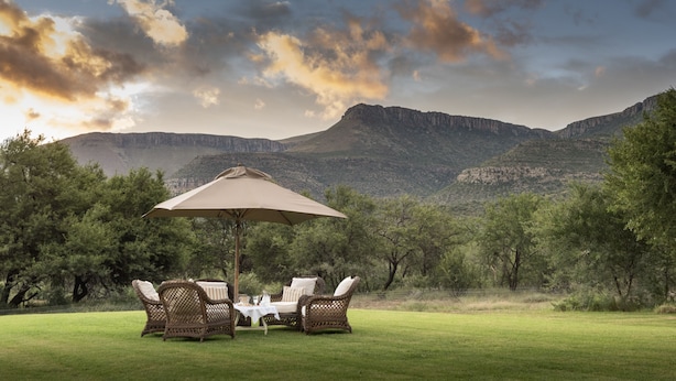 A patio with wicker furniture and a table under a large umbrella, overlooking a mountainous landscape. - Mount Camdeboo Private Game Reserve by NEWMARK (Blue Crane Route)