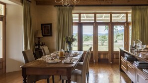 A dining area with a wooden table set for four, surrounded by chairs. A vase with flowers is on the table, and there are wine bottles and glasses. The room has wooden floors and a view through large glass doors to the outdoors. - Mount Camdeboo Private Game Reserve by NEWMARK (Blue Crane Route)