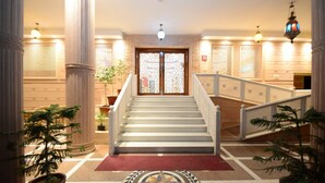 A grand staircase leading to a door with intricate glass panels, flanked by columns and potted plants, in a well-lit lobby area. - Lilypool - The Heritage Jalmahal (Jaipur)