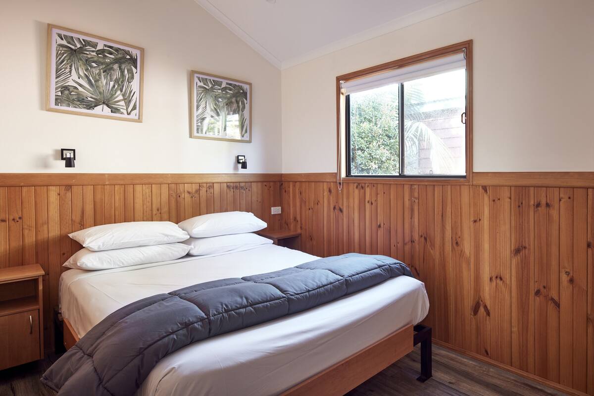 A bedroom with a wooden headboard, a large bed with white and grey bedding, two wall-mounted nightstands, and two framed tropical leaf paintings.