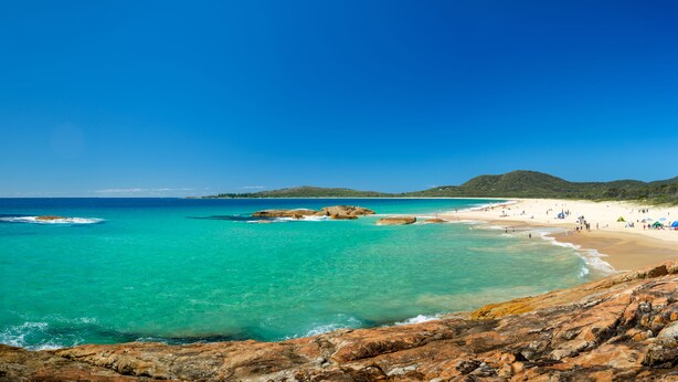 Une plage aux eaux turquoise et limpides, parsemée de rochers et bordée d’une étendue de sable où se trouvent des personnes et des tentes.