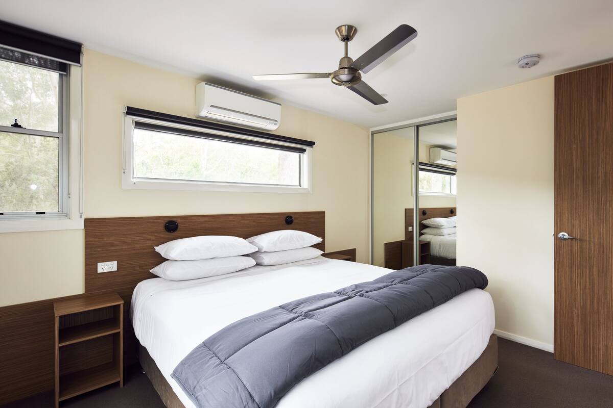 A neatly arranged bedroom with a bed, bedside tables, a ceiling fan, an air conditioning unit, and a window.
