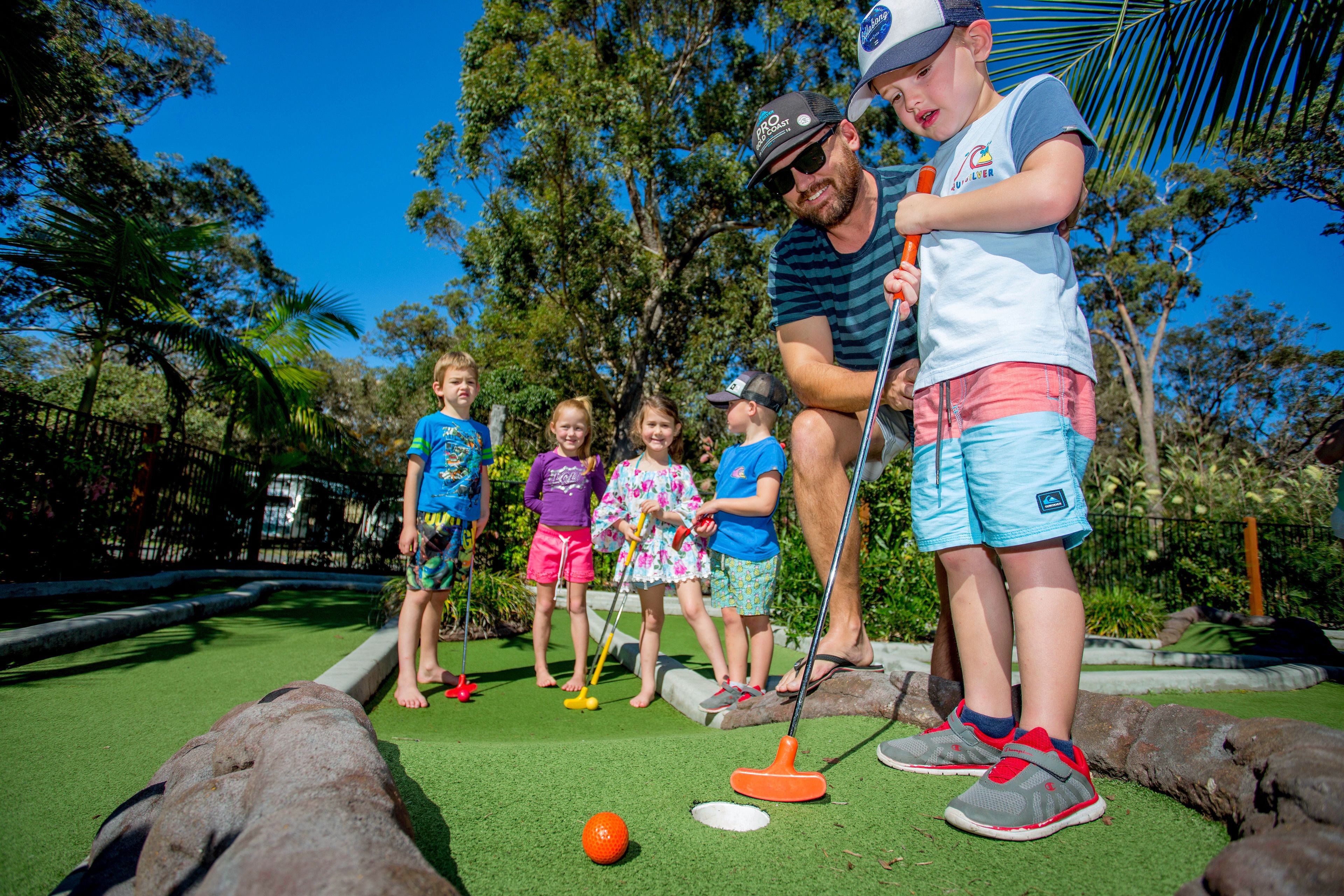 A group of people playing mini-golf on a green artificial turf with a clear blue sky in the background.