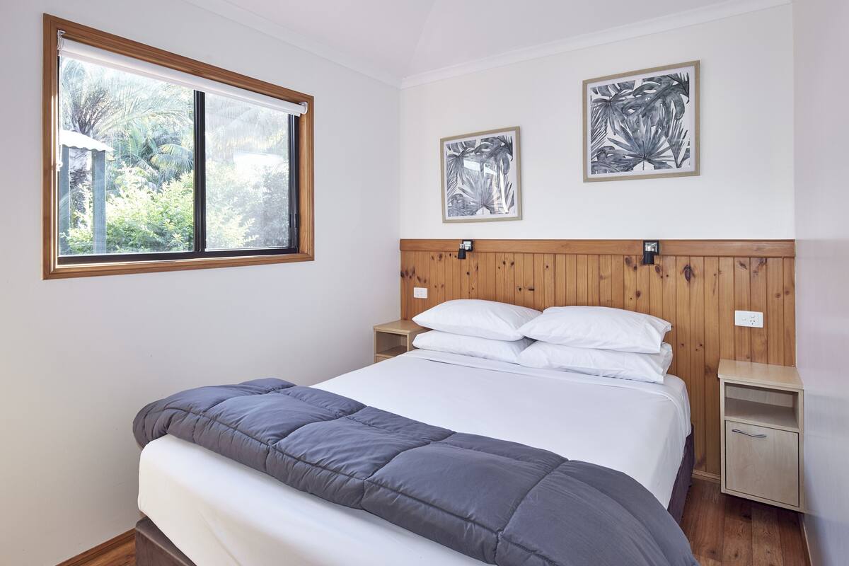 A bedroom with a bed, wooden headboard, two nightstands, a window with a view of greenery, and two framed pictures on the wall.