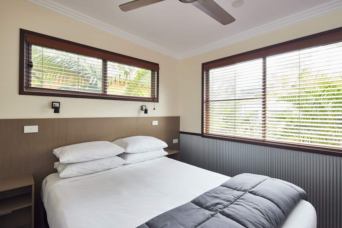 A neatly made bed with white and gray bedding, a wooden headboard, and a window with blinds.