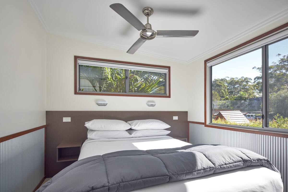 A bedroom with a bed, a ceiling fan, a window with a view of greenery, and a small shelf.