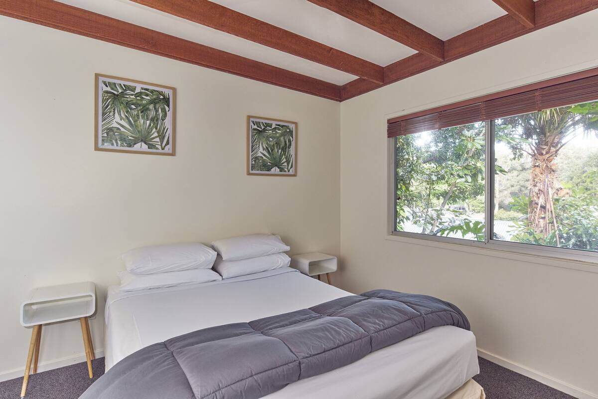 A bedroom with a bed, two wall-mounted picture frames, a bedside table, and a window with a view of greenery.