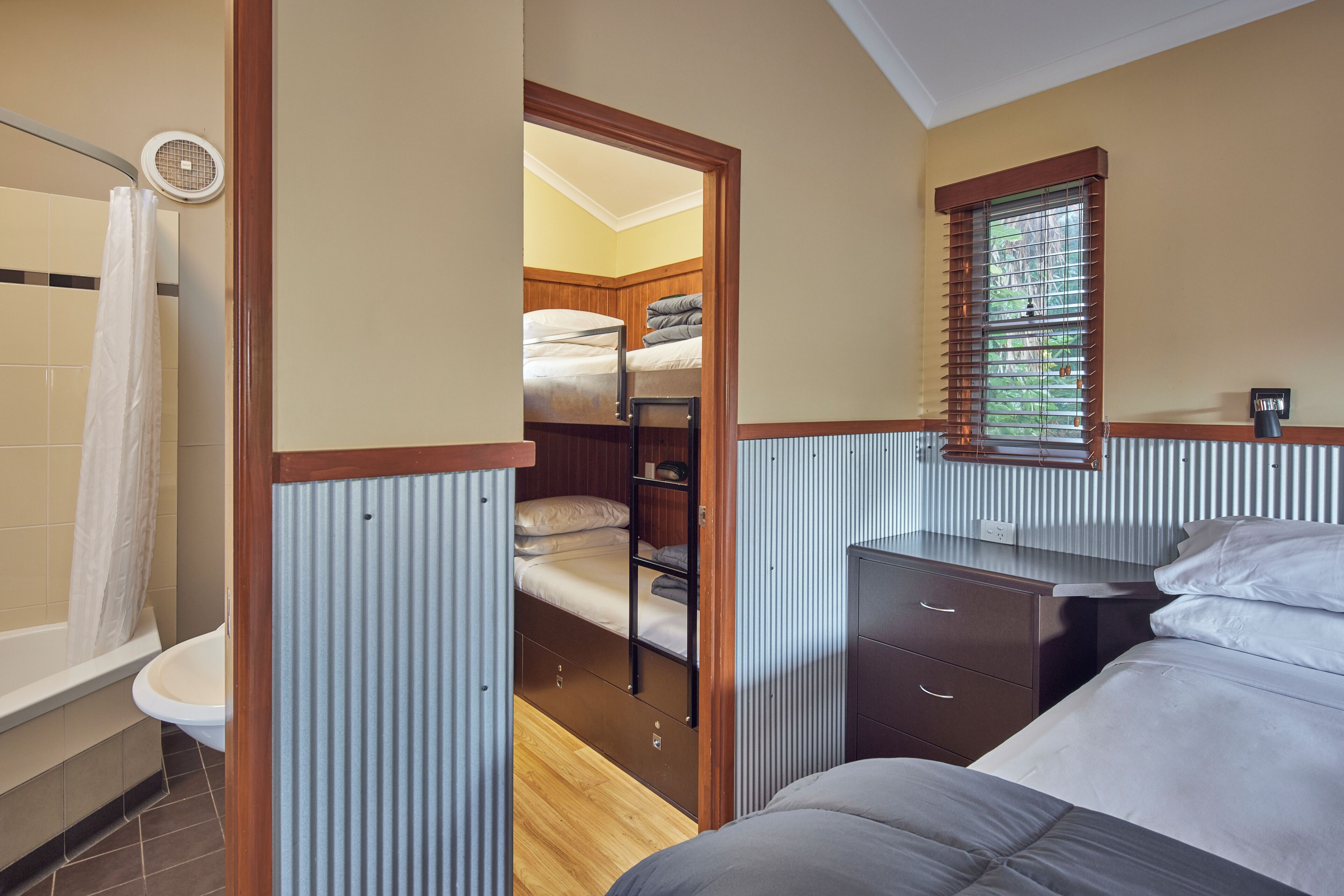 A compact bedroom with a bunk bed, a bedside table, a window with blinds, and a shower area.