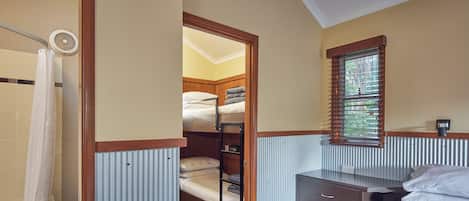A compact bedroom with a bunk bed, a bedside table, a window with blinds, and a shower area.