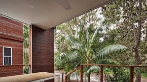 Une terrasse en bois, dotée d’un banc et d’une balustrade, qui offre une vue sur un jardin luxuriant, avec des palmiers et un bâtiment en bois.