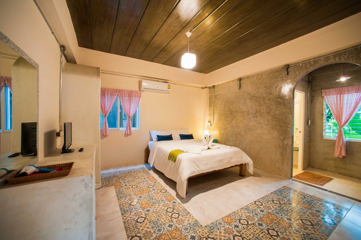 Un dormitorio con una cama, televisión, ventana con cortinas y suelo estampado.