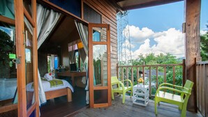 A wooden cabin with a balcony, a bed with a white blanket, a green chair, and a white table. - Baan Chokdee Pai Resort (Pai)