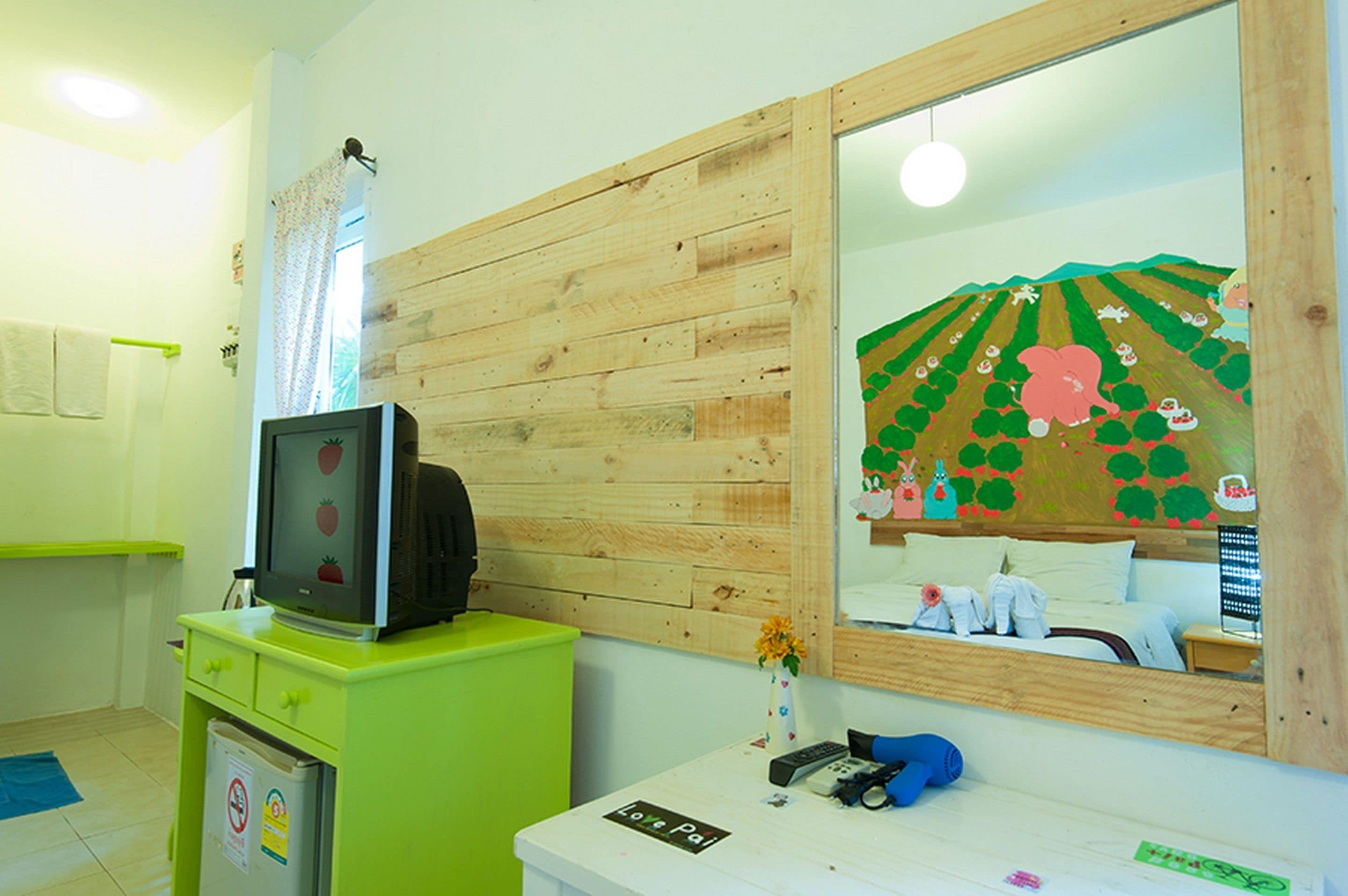 Una habitación con pared de madera, un espejo que refleja un paisaje caricaturesco, un televisor sobre un mueble verde y un escritorio pequeño con un cartel.