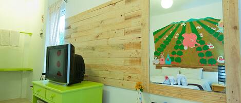 A room with a wooden wall, a mirror reflecting a cartoon landscape, a television on a green cabinet, and a small desk with a sign.