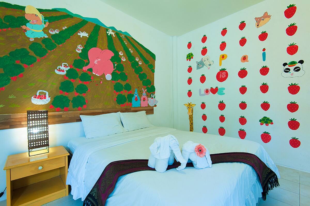 Un dormitorio con un mural grande en la pared, una cama con ropa de cama blanca, una mesita de noche y una pared decorada con pegatinas de fresas.