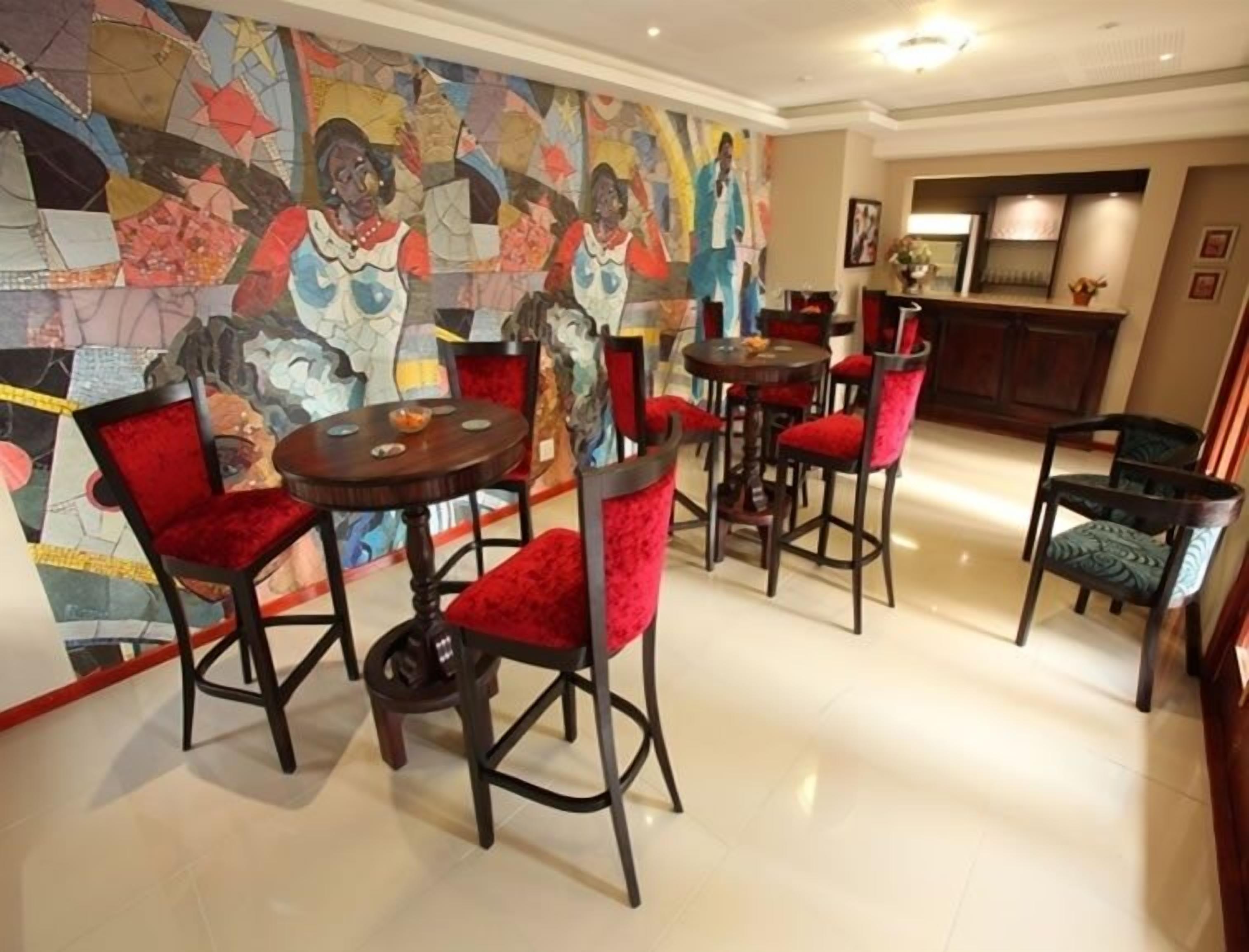A dining area with red upholstered chairs and round tables, a colorful abstract mural on the wall, and a bar area in the background.