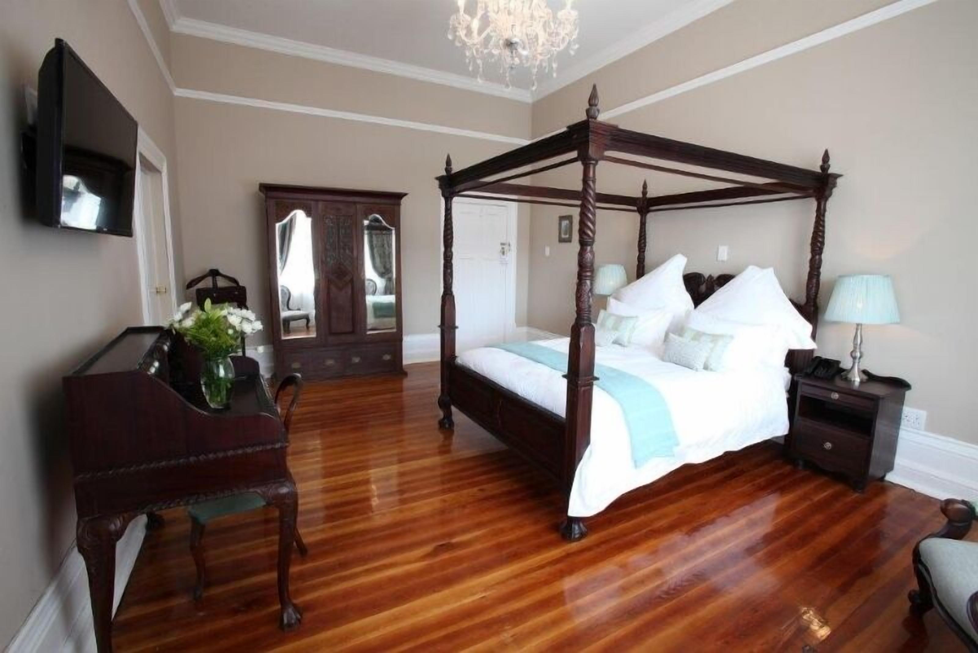 A four-poster bed with a wooden frame, a desk with a chair, a mirror, and a chandelier.