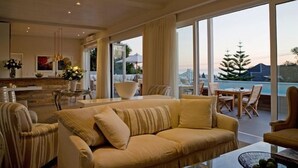 A spacious living room with a large sofa, a dining area, and a view of a swimming pool and greenery. - Sea Five Boutique Hotel (Cape Town)